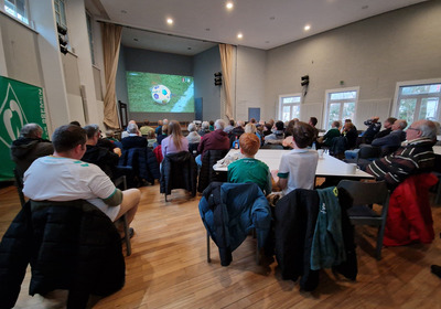 Eine Gruppe von Menschen sitzt in einem Veranstaltungssaal und schaut auf eine große Leinwand, auf der ein Fußballspiel übertragen wird. Die Zuschauer sind aufgeregt und tragen verschiedene Fanbekleidungen. Es sind Tische und Stühle sichtbar, und die Atmosphäre ist gesellig.