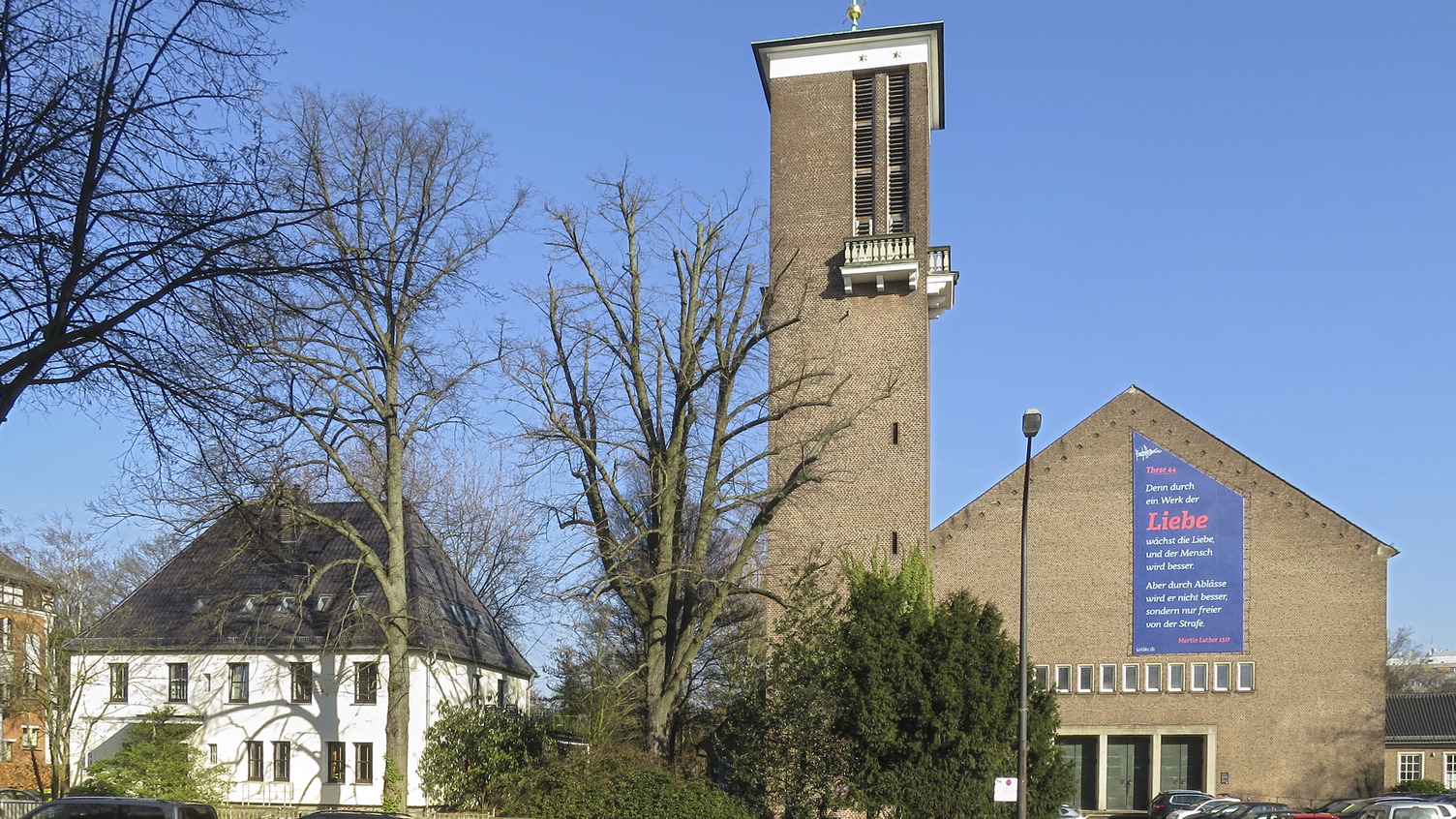 Das Bild zeigt eine Kirche mit einem hohen Turm und einem großen, bunten Banner, das eine Botschaft über Liebe enthält. Im Vordergrund stehen mehrere Autos, und im Hintergrund sind Bäume und ein weiteres Gebäude zu sehen. Der Himmel ist blau und klar.