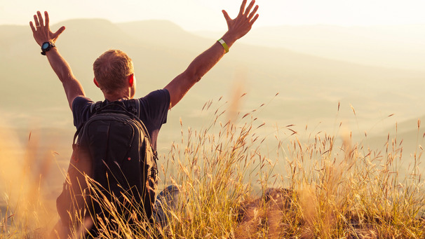 Eine Person mit einem Rucksack sitzt auf einem Hügel, hebt die Arme in die Luft und genießt die Aussicht auf eine weite, offene Landschaft bei Sonnenuntergang. Sträucher und Gräser umgeben die Person.