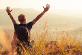 Eine Person mit einem Rucksack sitzt auf einem Hügel, hebt die Arme in die Luft und genießt die Aussicht auf eine weite, offene Landschaft bei Sonnenuntergang. Sträucher und Gräser umgeben die Person.