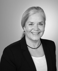 Porträt einer lächelnden Frau mit grauen Haaren in einem schwarzen Blazer. Sie hat eine helle Bluse und trägt eine Halskette. Der Hintergrund ist in sanften Grautönen gehalten.