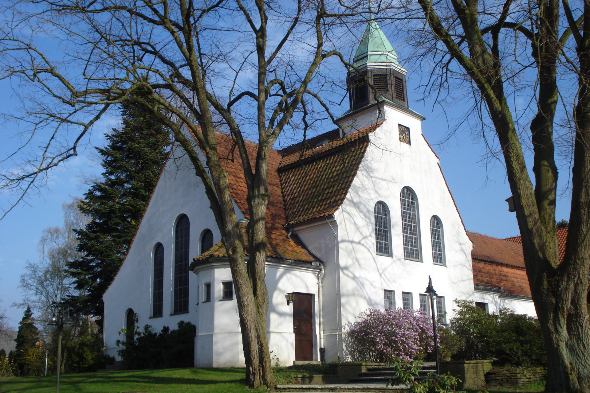 Ein helles Kirchengebäude mit einem spitzen Dach und einem kleinen Turm steht zwischen hohen Bäumen. Die Fenster sind groß und rundbogenförmig. Vor der Kirche blühen Pflanzen, und der Himmel ist klar und blau.