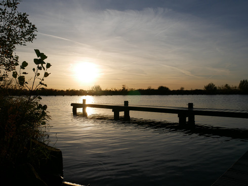 Ein Steg ragt hinaus aufs Wasser. Im Hintergrund geht die Sonne unter und wirft starke Schatten und Lichtreflektionen.