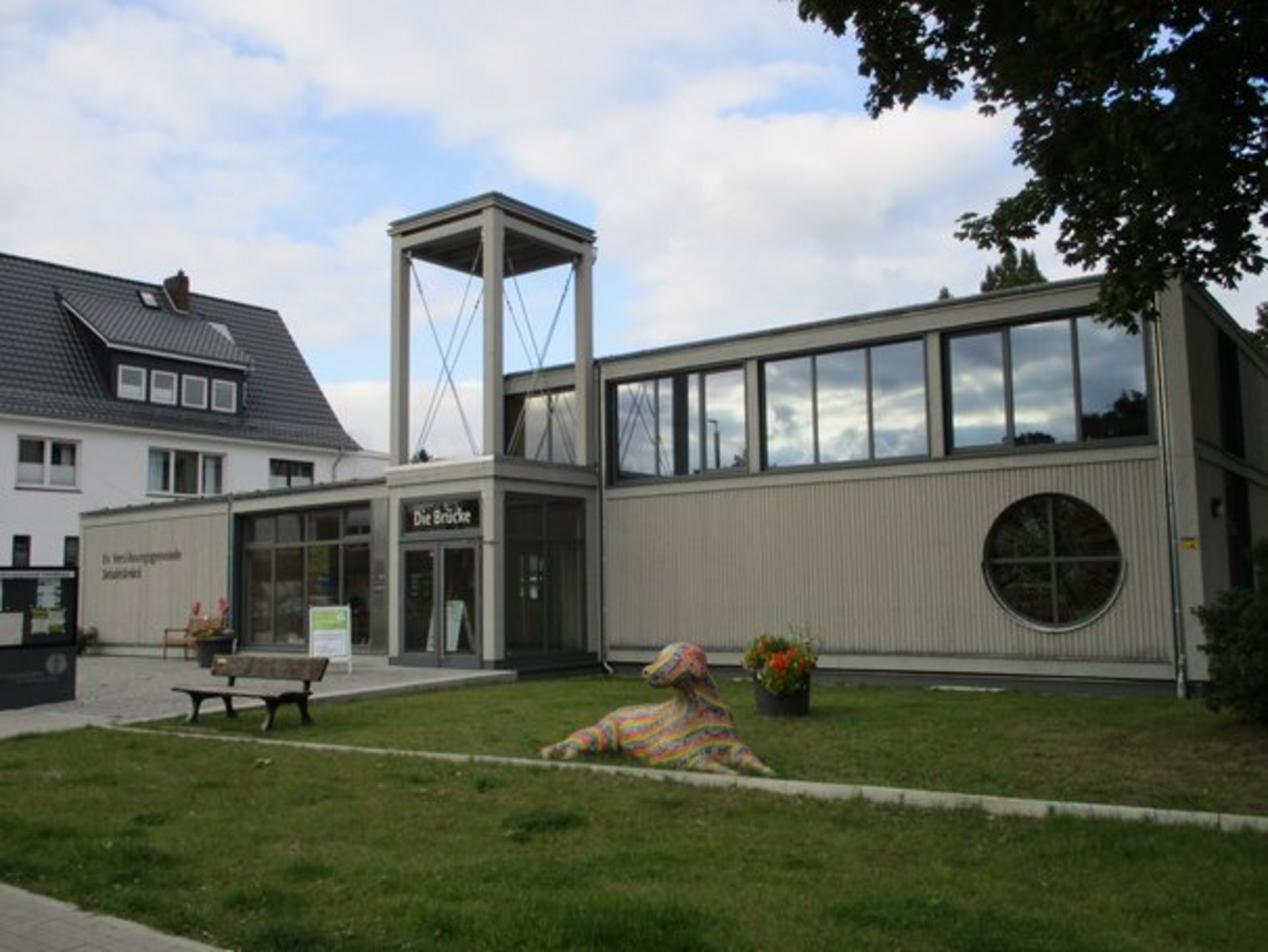 Ein modernes, zweigeschossiges Gebäude mit großen Fenstern und einem markanten Eingangsschild „Die Brücke“. Vor dem Gebäude befindet sich eine bunte, hundeartige Skulptur auf einer Rasenfläche. Im Hintergrund sind weitere Wohngebäude und Bäume sichtbar.