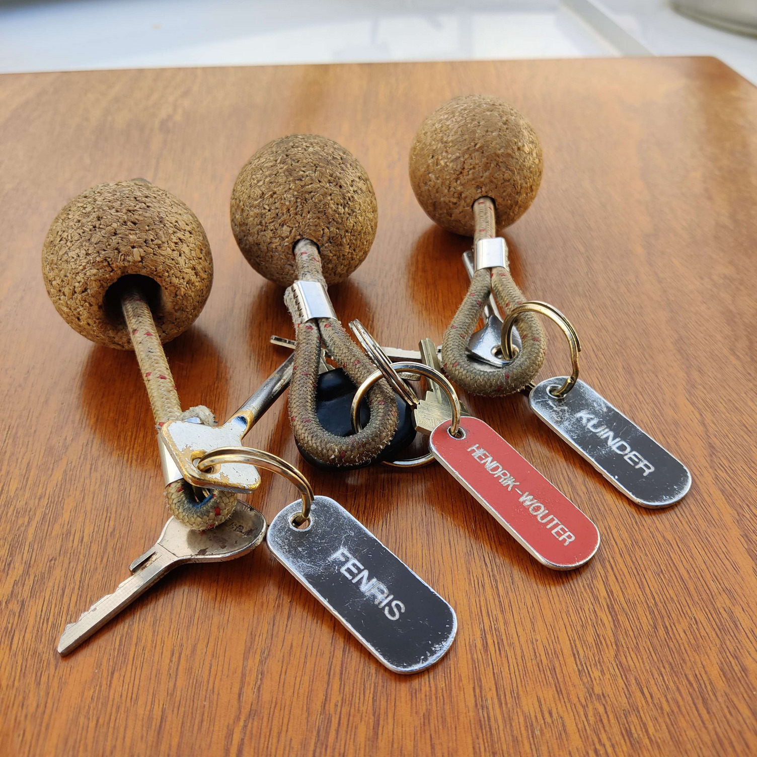 Bootsschlüssel Drei Schlüssel mit Korkkugeln und Schlüsselanhängern. Die Anhänger sind mit den Worten "Fernis", "Hendrik Wouter" und "Kuider" beschriftet. Der Hintergrund ist eine braune Holzoberfläche.