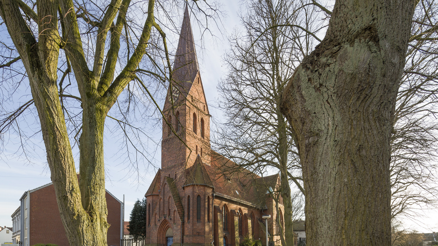 Eine rote Backsteinkirche mit einem hohen, spitzen Turm ist im Bild zu sehen. Sie ist umgeben von Bäumen und Rasen. Im Hintergrund sind einige kleinere Gebäude sichtbar.