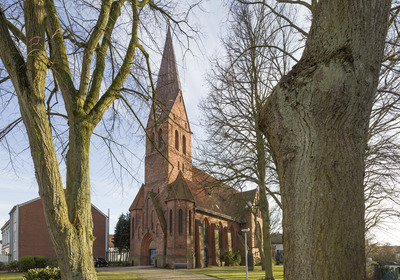 Eine rote Backsteinkirche mit einem hohen, spitzen Turm ist im Bild zu sehen. Sie ist umgeben von Bäumen und Rasen. Im Hintergrund sind einige kleinere Gebäude sichtbar.