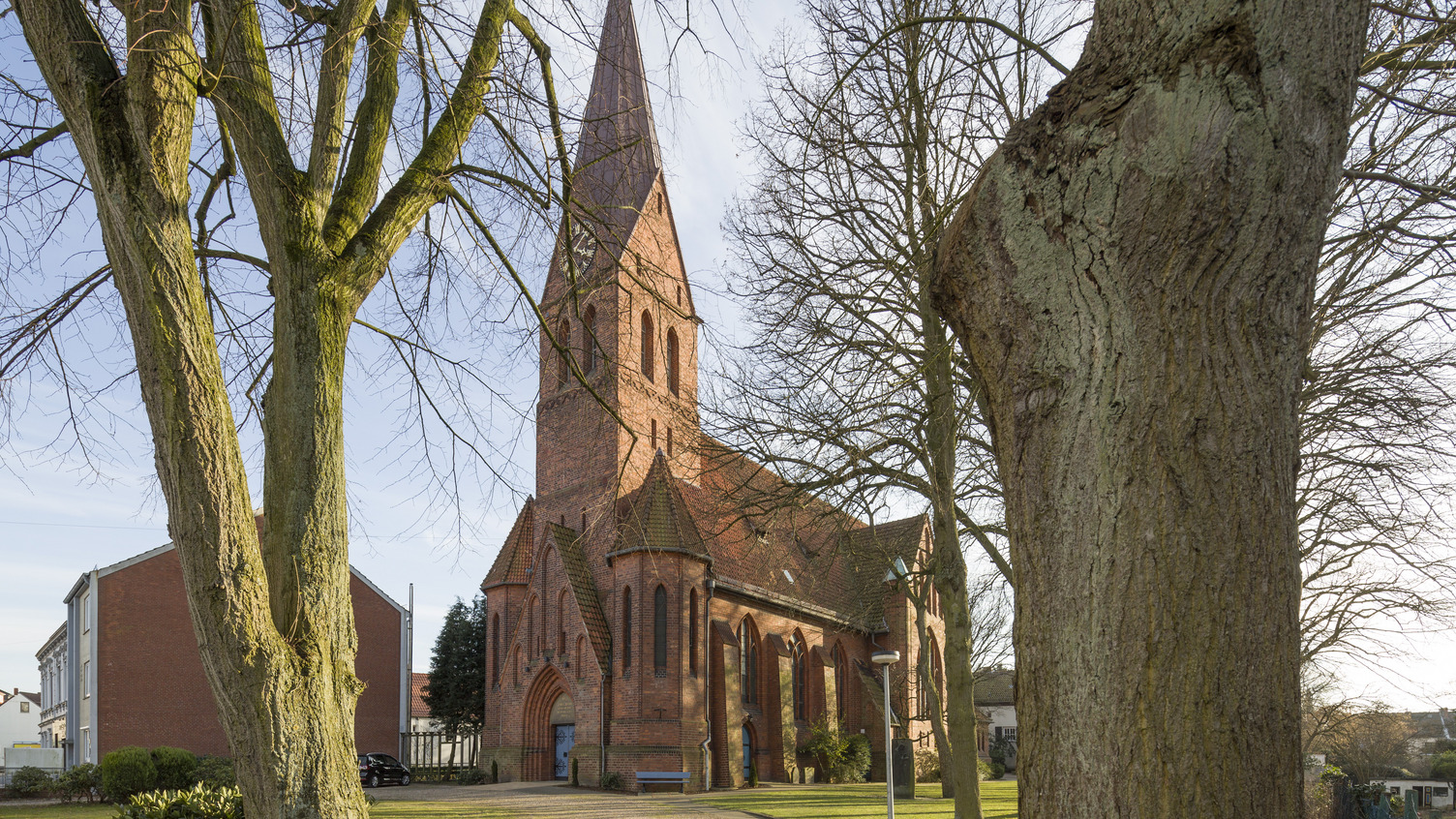 Eine rote Backsteinkirche mit einem hohen, spitzen Turm ist im Bild zu sehen. Sie ist umgeben von Bäumen und Rasen. Im Hintergrund sind einige kleinere Gebäude sichtbar.