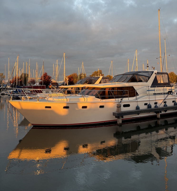 Sloten am Abend Ein weißes Boot liegt im Hafen, vorne hell erleuchtet von der Abendsonne. Es spiegelt sich im ruhigen Wasser, umgeben von mehreren weiteren Booten und Masten. Der Himmel ist mit sanften, grauen Wolken durchzogen, die das Licht der untergehenden Sonne reflektieren.