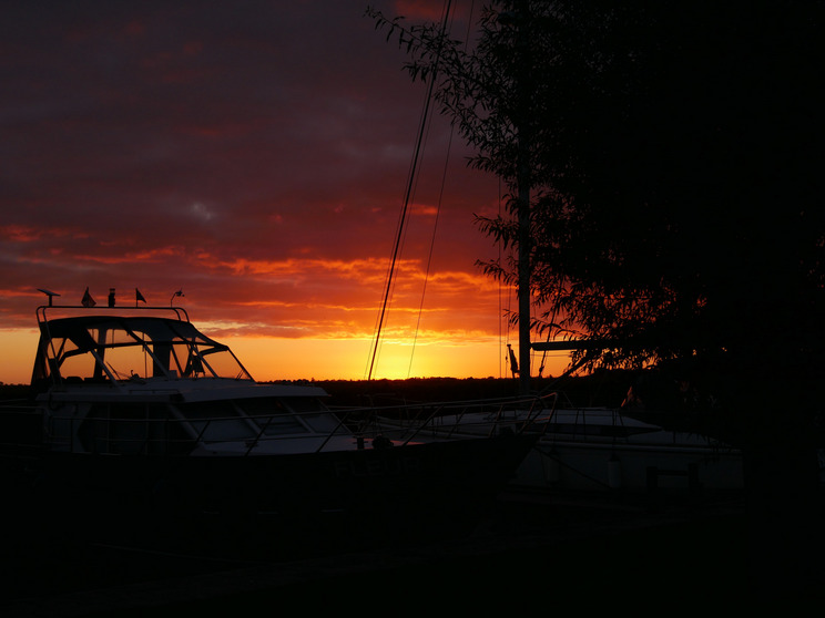 Sloten Sonnenuntergang Ein Boot liegt am Ufer, silhouettiert vor einem strahlenden Sonnenuntergang. Der Himmel zeigt kräftige orange und rote Farben, während sich einige Wolken daran vorbeiziehen. Die Szenerie vermittelt eine ruhige und friedliche Atmosphäre.