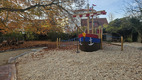 Ein Spielplatz mit einem großen, bunten Holzschiff als Spielgerät. Das Schiff hat rote und blaue Streifen und Anker-Details. Umgeben von einem schottrigen Boden und herabfallenden, orangefarbenen Blättern der Bäume, vermittelt es eine einladende und spielerische Atmosphäre.
