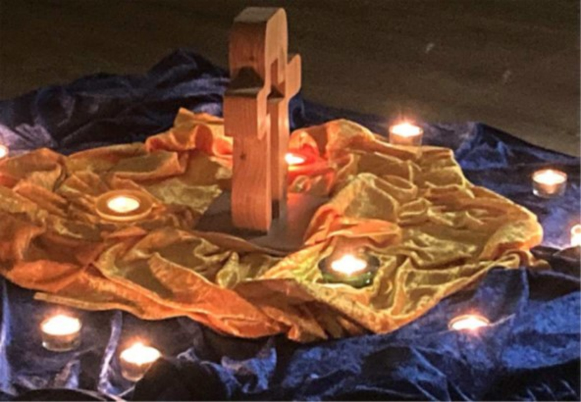 Ein Holzaltar mit einem Kreuz steht auf einer gelben Stoffunterlage, umgeben von schwach leuchtenden Kerzen. Der Hintergrund besteht aus blauen und goldenen Stoffen, die dem Arrangement eine ruhige, spirituelle Atmosphäre verleihen.