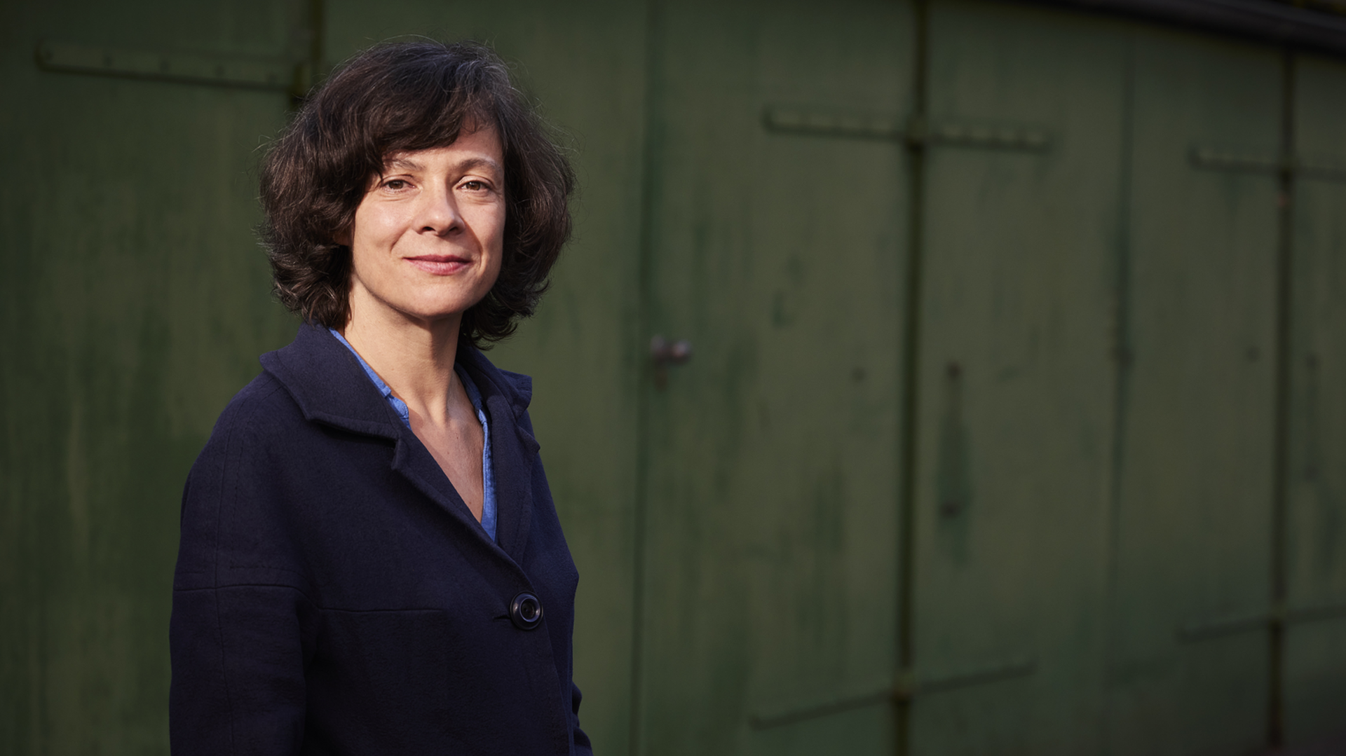 Porträt einer Frau mit lockigem, dunklem Haar, die in einem blauen Mantel lächelt. Der Hintergrund zeigt eine grüne Wand mit geschlossenen Türen. Die Frau steht in einer aufrechten Position und strahlt Selbstbewusstsein aus.