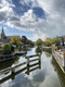 Der Blick führt auf einer Brücke einen Kanal hinunter der durch den Ort fließt. Die Wolken am blauen Himmel spiegeln sich auf der Wasseroberfläche.
