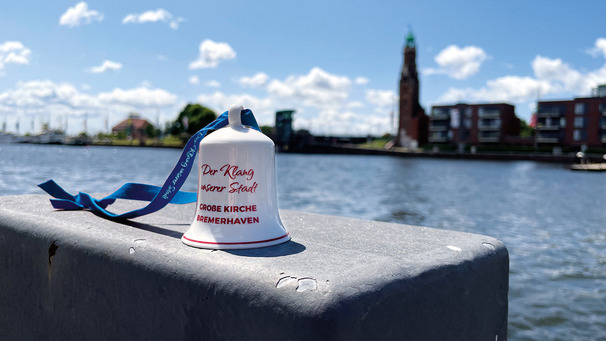 Eine kleine, weiße Glocke mit rotem Text steht auf einer grauen Oberfläche. Der Text lautet „Der Klang unserer Stadt“ und darunter „GROßE KIRCHE BREMENHAVEN“. Im Hintergrund sieht man den Wasserlauf und einen Kirchturm sowie einige Gebäude. Der Himmel ist blau mit vielen Wolken.