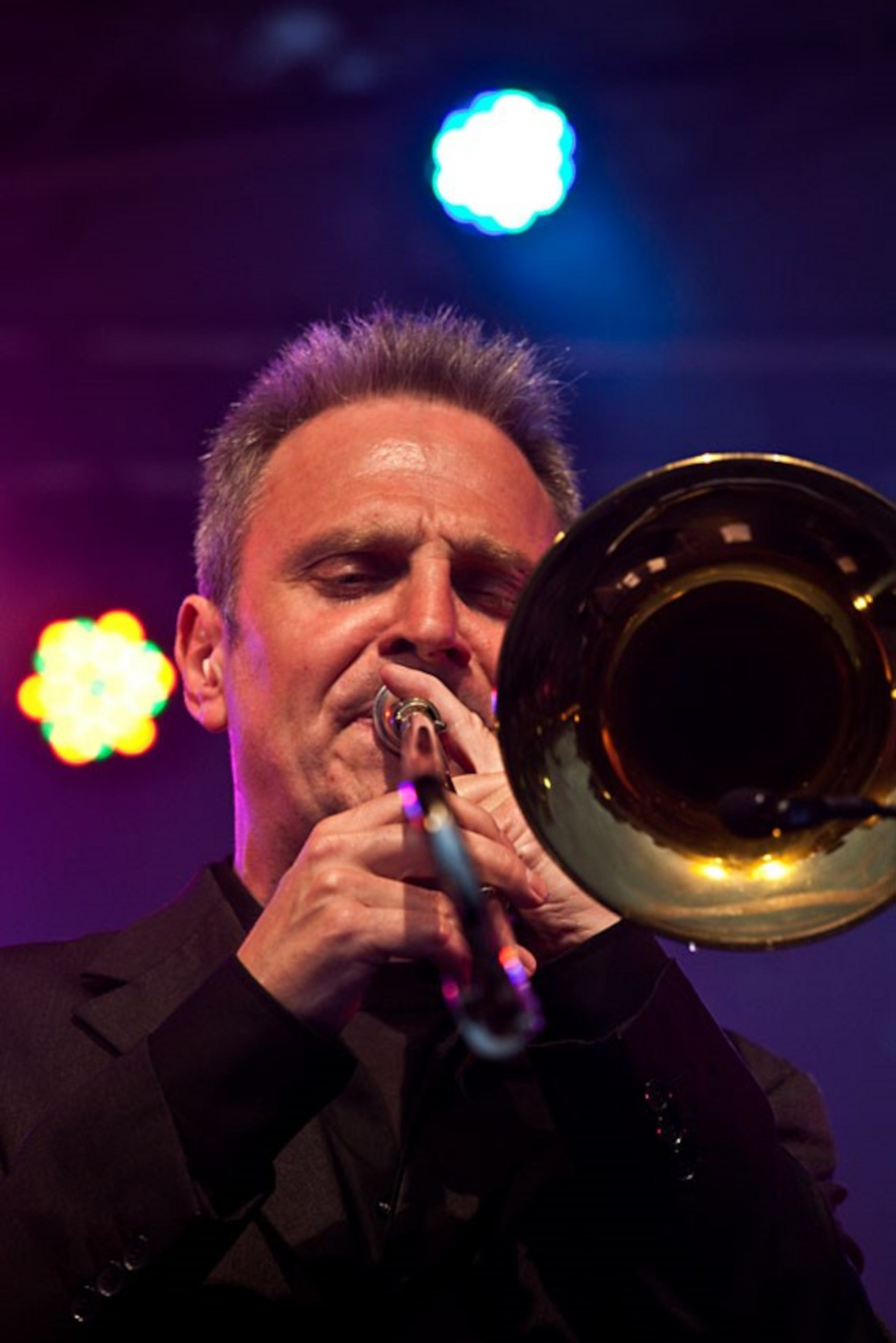 Ein Musiker spielt Trompete auf einer Bühne. Er trägt ein schwarzes Outfit und konzentriert sich intensiv auf die Musik. Im Hintergrund sind bunte Lichter zu sehen, die zur Stimmung des Auftritts beitragen.