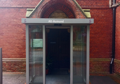 Eingang zu einem Gebäude mit dem Schild "Alt-Aumund". Die Eingangstür ist aus Glas und befindet sich in einer roten Backsteinfassade. Im Vordergrund steht eine Pflanze.