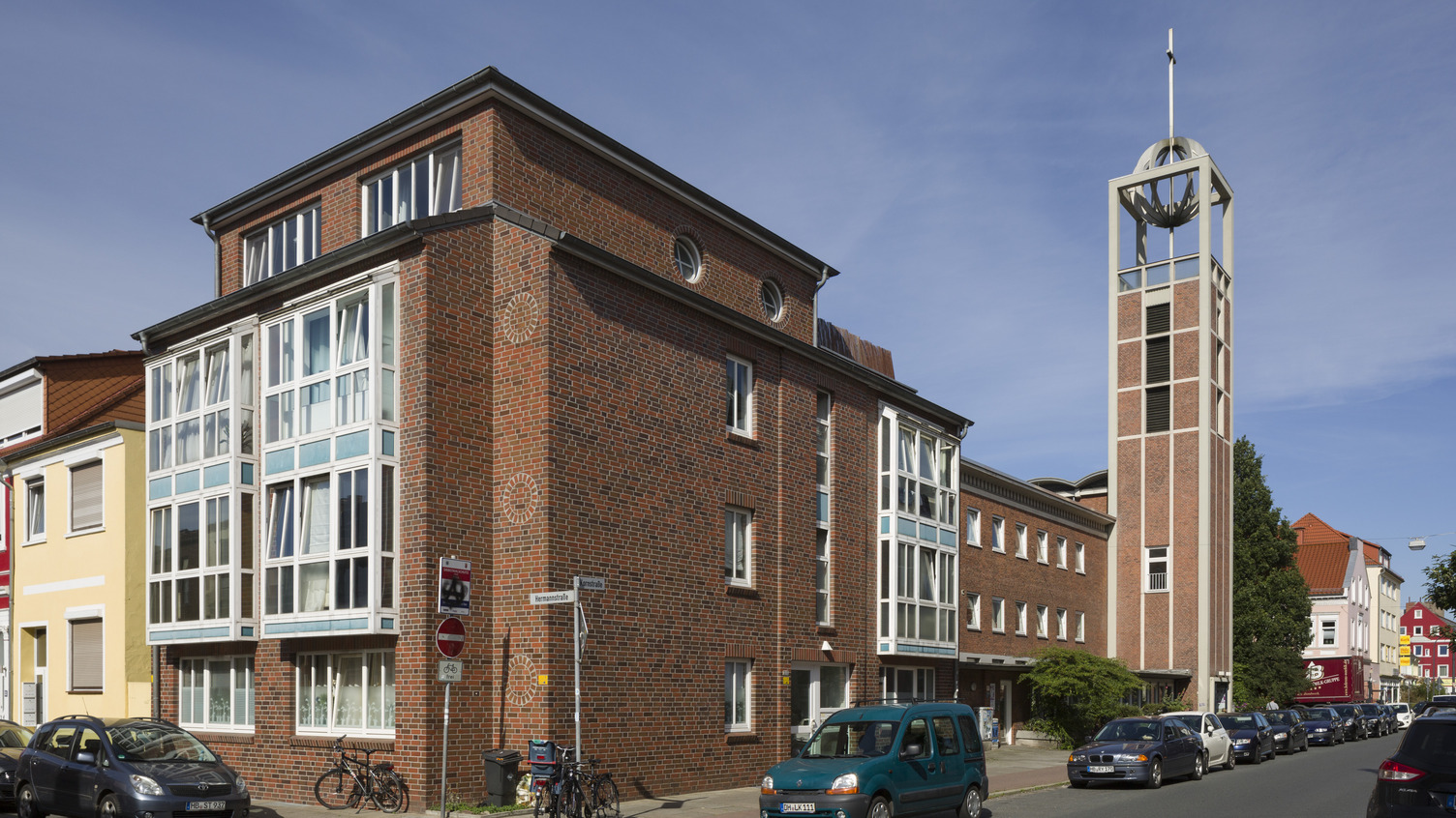 Ein rechteckiges, mehrstöckiges Gebäude aus rotem Backstein mit großen Fenstern und einem hohen, schmalen Glockenturm ist zu sehen. Der Turm hat ein markantes Design und eine Kugelspitze. Auf der Straße parken mehrere Autos, und es gibt Fahrradständer. Der Himmel ist klar und blau.