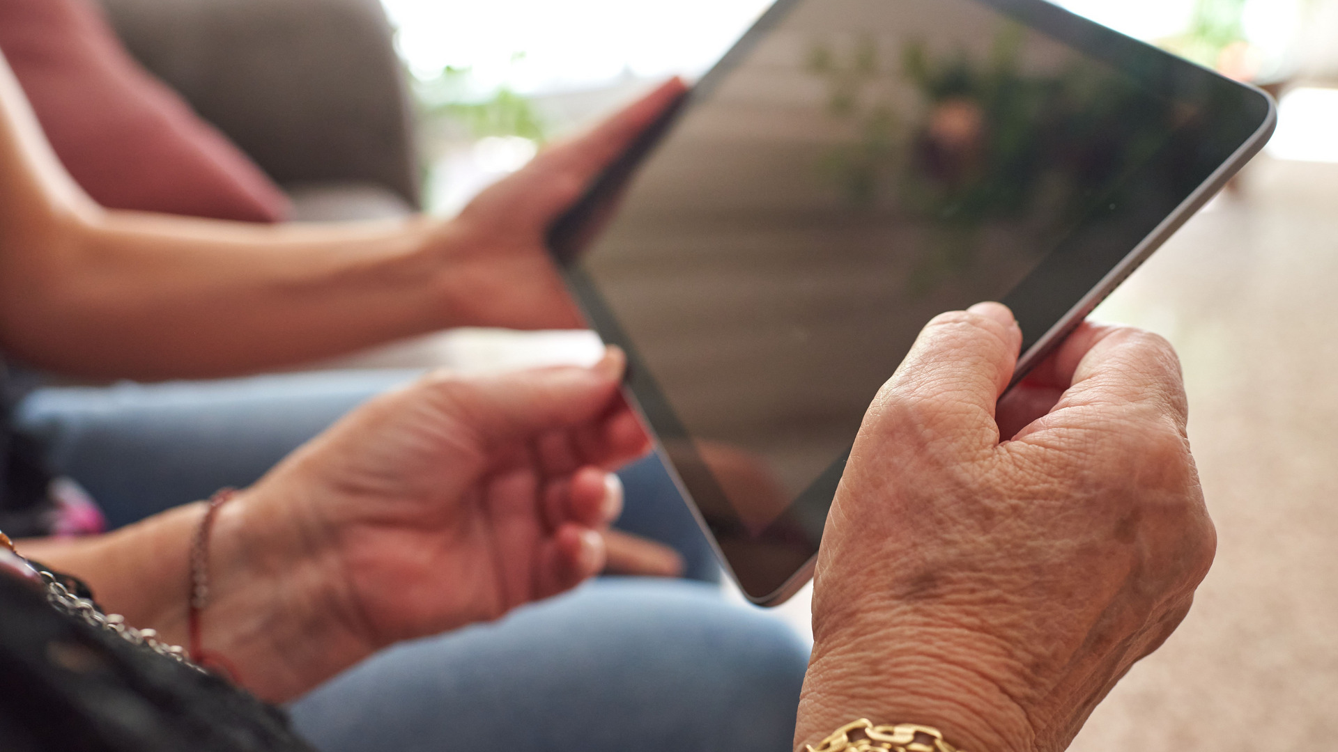 Eine Person hält ein Tablet mit beiden Händen, während sie auf den Bildschirm schaut. Die Umgebung scheint gemütlich und wohnlich zu sein, mit unscharfen Hinweisen auf Möbel im Hintergrund. Die Hände sind unterschiedlich alt und zeigen Details wie eine goldene Armbanduhr.