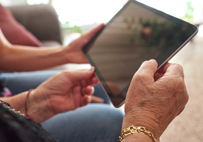 Eine Person hält ein Tablet mit beiden Händen, während sie auf den Bildschirm schaut. Die Umgebung scheint gemütlich und wohnlich zu sein, mit unscharfen Hinweisen auf Möbel im Hintergrund. Die Hände sind unterschiedlich alt und zeigen Details wie eine goldene Armbanduhr.