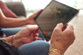 Eine Person hält ein Tablet mit beiden Händen, während sie auf den Bildschirm schaut. Die Umgebung scheint gemütlich und wohnlich zu sein, mit unscharfen Hinweisen auf Möbel im Hintergrund. Die Hände sind unterschiedlich alt und zeigen Details wie eine goldene Armbanduhr.