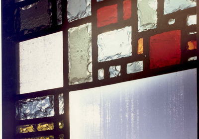 Ein farbiges Glasfenster mit geometrischem Muster, das verschiedene Farbtöne wie Rot, Grün, Gelb und Blau enthält. Die einzelnen Glasstücke sind von schwarzem Glasrahmen umgeben und weisen unterschiedliche Texturen auf, wodurch Licht durchgelassen wird und ein spielerisches Lichtspiel entsteht.
