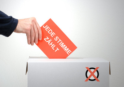 Die Illustration zeigt eine Hand, die einen Stimmzttel in eine Wahlurne wirft