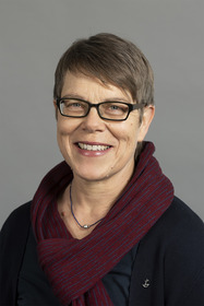 Porträt einer lächelnden Frau mit kurzen, braunen Haaren und einem roten Schal. Sie trägt eine dunkle Bluse und hat eine Brille auf. Der Hintergrund ist grau und neutral.