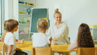 Drei Kinder sitzen mit dem Rücken zur Kamera an einem Tisch und gucken zu einer ebenfalls am Tisch sitzenden Frau.