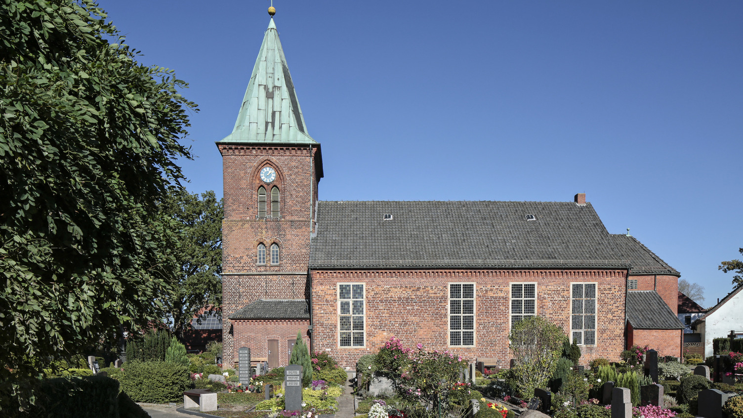 Eine rote Backsteinkirche mit einem höheren, grünen Turm. Der Kirchhof ist mit Blumen und Gräbern geschmückt. Im Hintergrund ist ein blauer Himmel zu sehen.