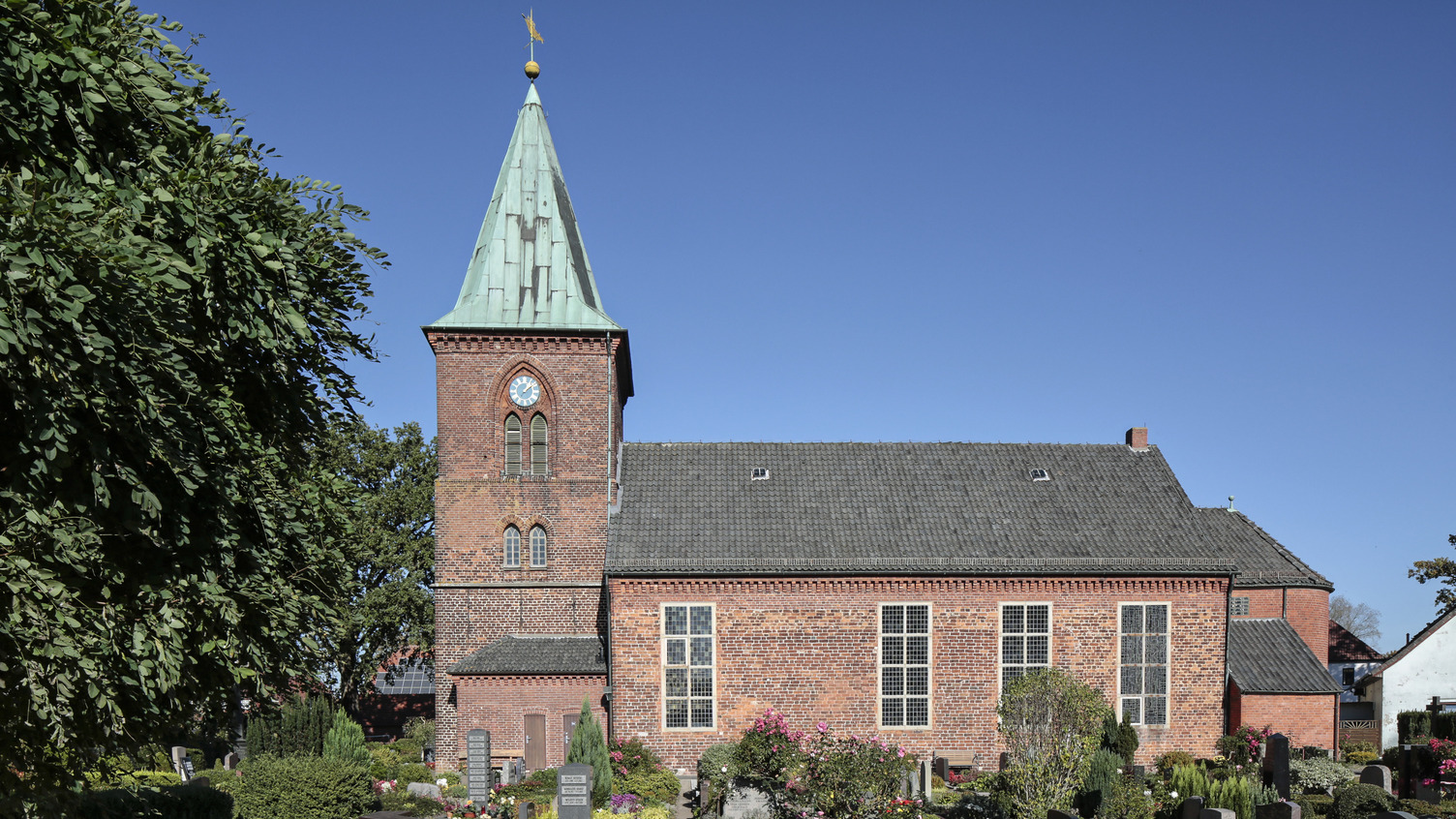 Eine rote Backsteinkirche mit einem höheren, grünen Turm. Der Kirchhof ist mit Blumen und Gräbern geschmückt. Im Hintergrund ist ein blauer Himmel zu sehen.