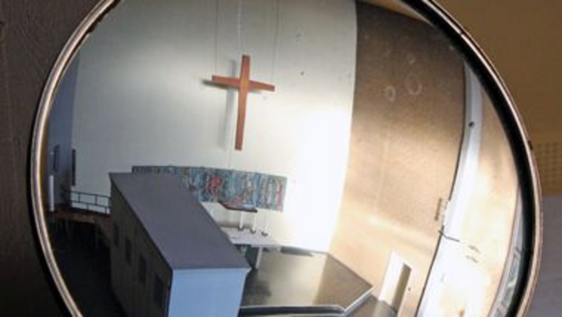 In einem runden Spiegel ist eine Kirche zu sehen. Der Hauptfokus liegt auf einem Holz-Kreuz, das über dem Altar schwebt, in einem hell erleuchteten Raum. Die Wand ist mit einem bunten Gemälde dekoriert.