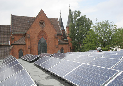 Solarzellen werden auf einem Flachdach installiert. Im Hintergrund ist die Friedenskirche zu sehen.