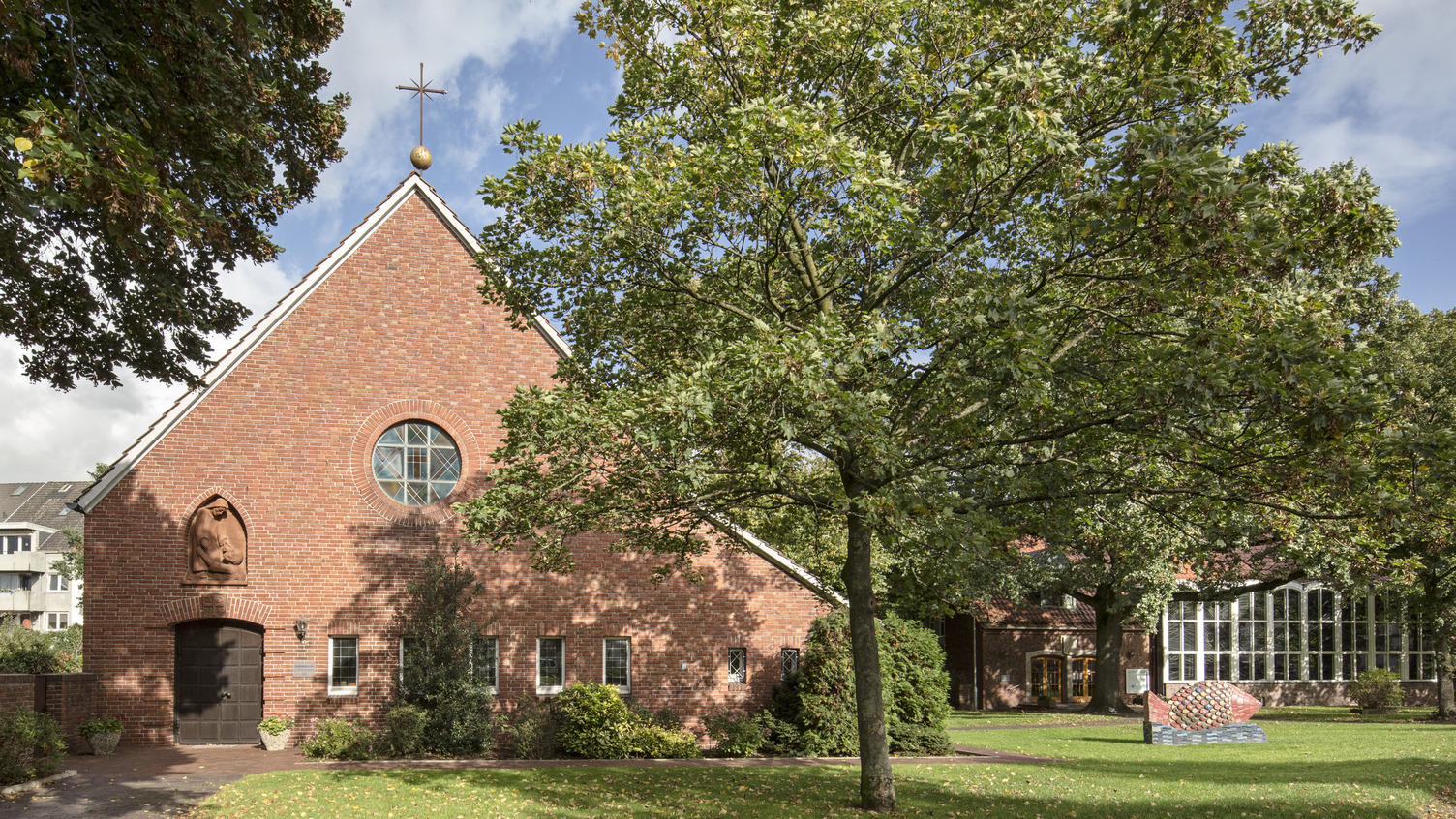 Ein rotes Backsteingebäude mit einem spitzen Dach und einem Kreuz an der Spitze. Vor dem Gebäude stehen Bäume und einige Pflanzen. 