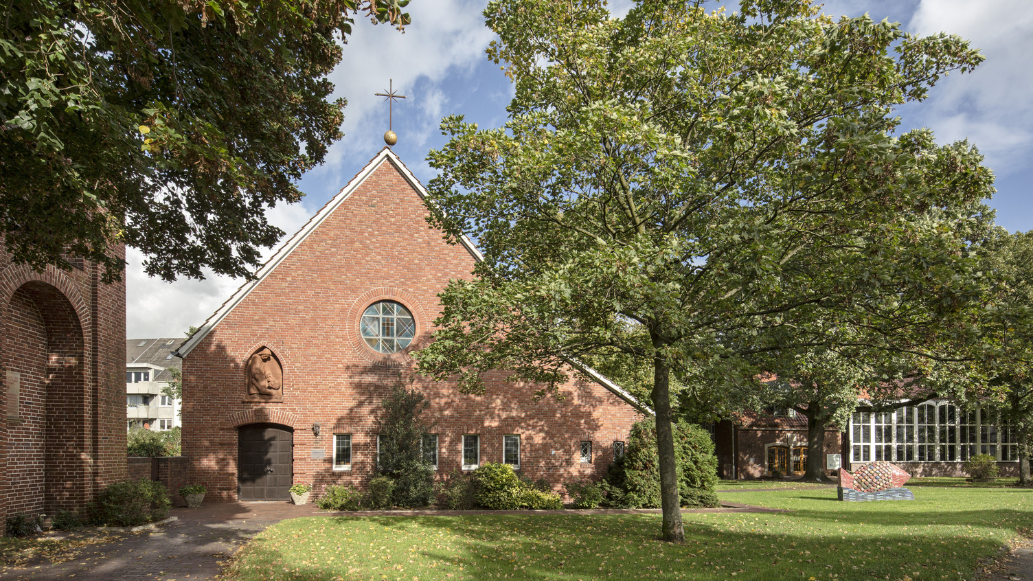 Ein rotes Backsteingebäude mit einem spitzen Dach und einem Kreuz an der Spitze. Vor dem Gebäude stehen Bäume und einige Pflanzen. 