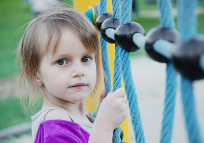 Ein kleines Mädchen mit langen, braunen Haaren und blauen Augen hält sich an einem Kletterseil fest. Sie trägt ein lila Oberteil und schaut in die Kamera. Der Hintergrund zeigt eine grüne Wiese und andere Kinder, die spielen.