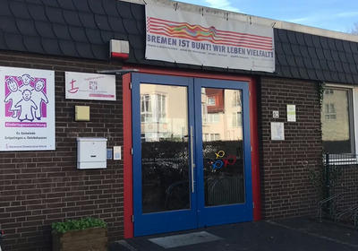 Das Bild zeigt den Eingang einer Kindertagesstätte in Bremen. Über der blauen Doppeltür hängt ein großes Banner mit dem Schriftzug „Bremen ist bunt! Wir leben Vielfalt!“ Neben der Tür befinden sich Informationen zur Einrichtung sowie ein Briefkasten.