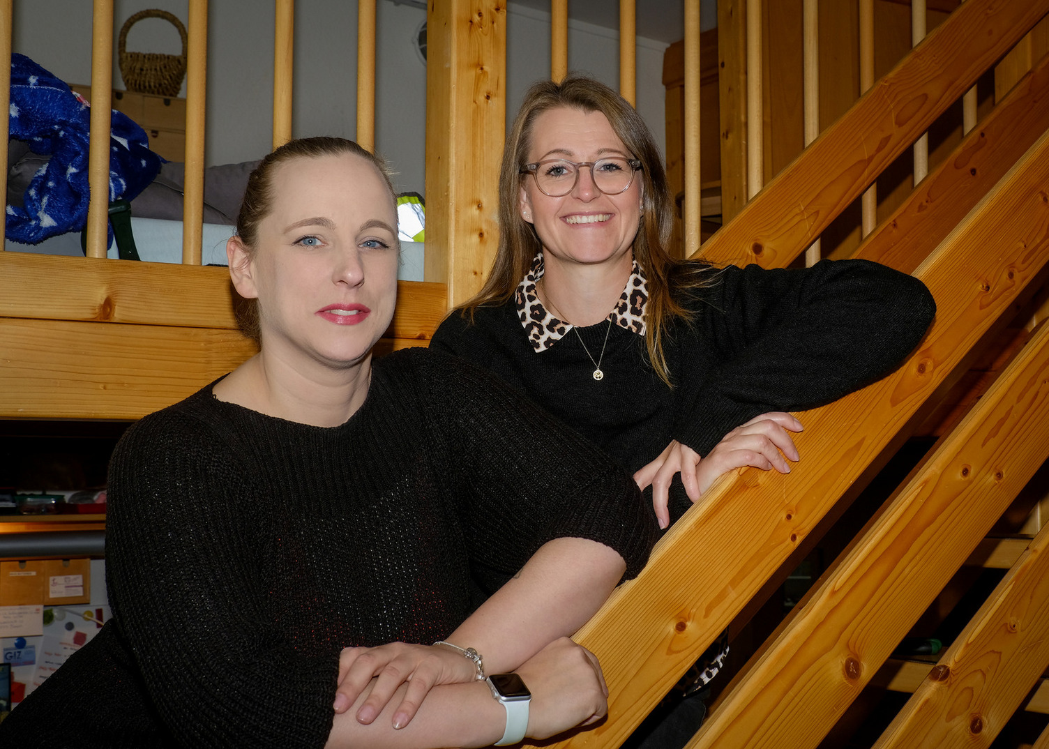 Zwei Frauen stehen lächelnd an einer Holztreppe. Eine trägt ein schwarzes Oberteil, die andere hat ein Oberteil mit einem Leopardenmuster. Die Umgebung wirkt gemütlich und hell, mit Holz- und Wohnaccessoires im Hintergrund.