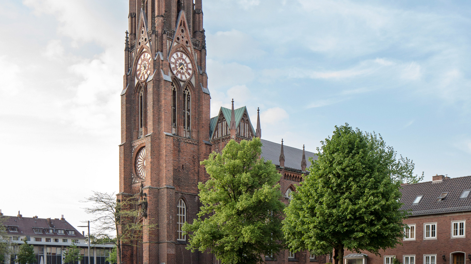 Ein historischer Glockenturm aus Backstein steht im Fokus des Bildes, umgeben von Bäumen. Der Turm hat auffällige Ziffernblätter und eine reich verzierte Fassade. Im Hintergrund sind weitere Gebäude und der Himmel sichtbar, der teilweise bewölkt ist.