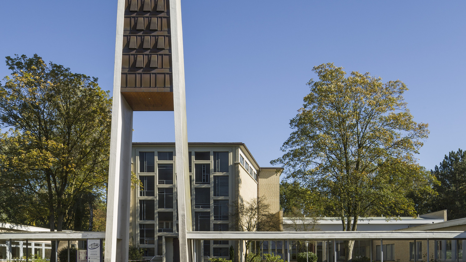 Ein moderner Bau mit einem hohen, schmalen Kirchturm steht im Vordergrund. Der Turm hat eine markante, geometrische Form und ist aus Beton mit Holzverkleidung. Im Hintergrund ist ein zweigeschossiges Gebäude mit Glasfenstern zu sehen, umgeben von Bäumen und einem Parkplatz.