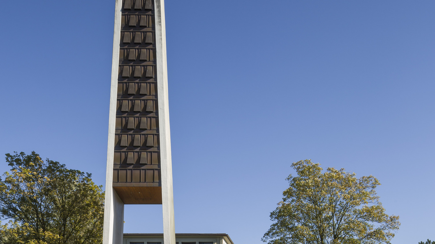 Ein moderner Bau mit einem hohen, schmalen Kirchturm steht im Vordergrund. Der Turm hat eine markante, geometrische Form und ist aus Beton mit Holzverkleidung. Im Hintergrund ist ein zweigeschossiges Gebäude mit Glasfenstern zu sehen, umgeben von Bäumen und einem Parkplatz.