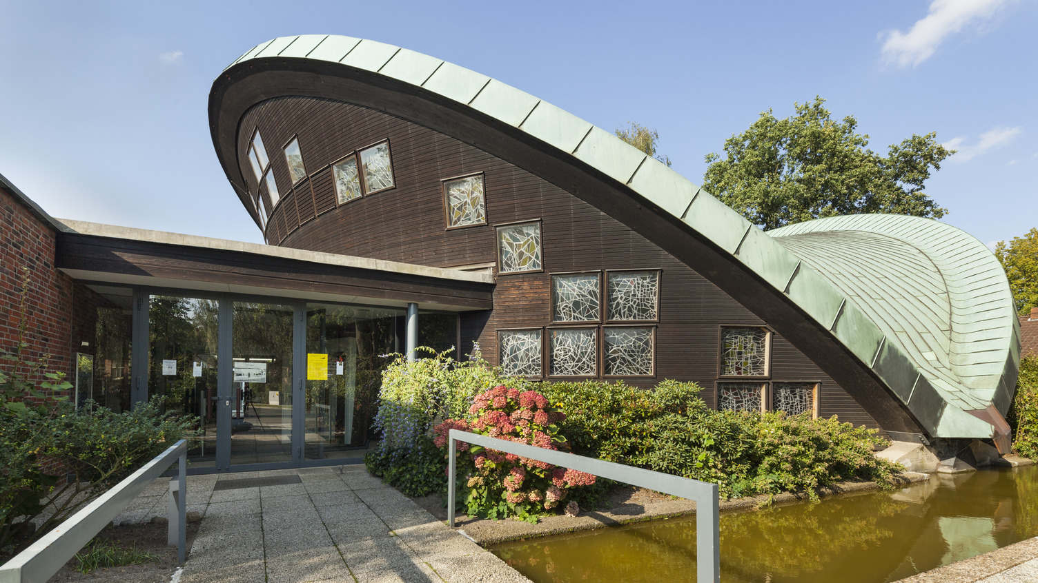 Ein modernes Gebäude mit einer markanten, geschwungenen Dachkonstruktion aus Metall und Holz. Die Fassade hat große Fenster mit geometrischen Mustern. Vor dem Eingang befindet sich ein gepflasterter Weg, umgeben von bunten Blumen und Grünpflanzen.