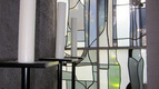 Im Vordergrund sind mehrere hohe, weiße Kerzen auf schwarzen Halterungen zu sehen. Im Hintergrund befindet sich ein buntes, modernes Fenster mit geometrischen Glasformen, das sanftes Licht durchlässt. Die Atmosphäre wirkt ruhig und spirituell.