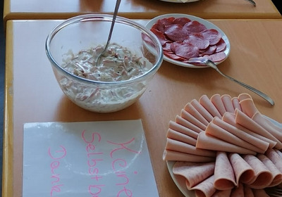 Ein gedeckter Tisch mit verschiedenen Lebensmitteln: gekochte Eier, Käse und Aufschnitt sind in Reihen angeordnet. Dazu gibt es eine Schüssel mit Salat, Marmelade und kleine Portionspackungen mit Butter. Ein Schild weist darauf hin, dass Selbstbedienung nicht erlaubt ist.