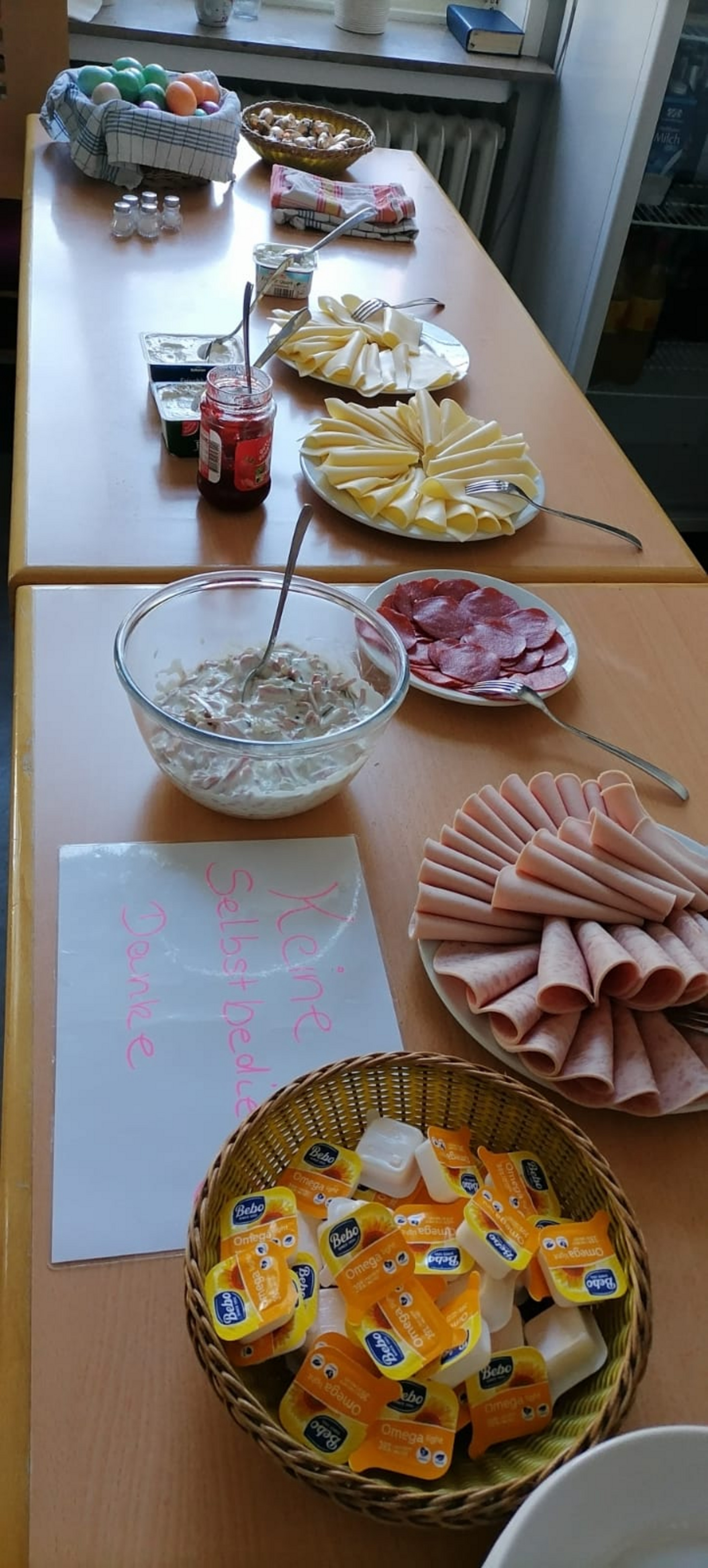 Ein gedeckter Tisch mit verschiedenen Lebensmitteln: gekochte Eier, Käse und Aufschnitt sind in Reihen angeordnet. Dazu gibt es eine Schüssel mit Salat, Marmelade und kleine Portionspackungen mit Butter. Ein Schild weist darauf hin, dass Selbstbedienung nicht erlaubt ist.