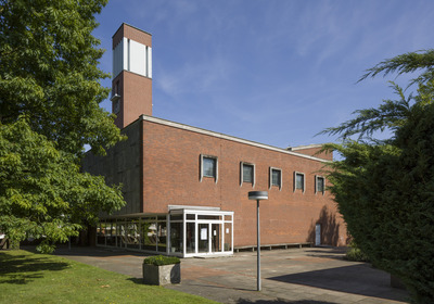 Ein modernes, zweigeschossiges Ziegelgebäude mit großen Fenstern und einem markanten, höheren Turm. Der Eingangsbereich ist durch ein transparentes Vordach geschützt. Um das Gebäude herum sind grüne Bäume und Sträucher sichtbar, bei klarem Himmel und Sonnenschein.
