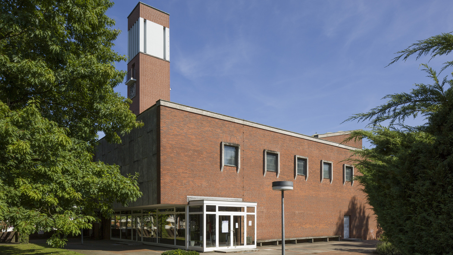 Ein modernes, zweigeschossiges Ziegelgebäude mit großen Fenstern und einem markanten, höheren Turm. Der Eingangsbereich ist durch ein transparentes Vordach geschützt. Um das Gebäude herum sind grüne Bäume und Sträucher sichtbar, bei klarem Himmel und Sonnenschein.