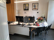 Ein Büro mit einem weißen Tisch, zwei Monitoren und Büromaterial. Im Hintergrund sind Holzschränke und ein Garderobenständer. Auf dem Tisch steht eine Blumenvase mit Blumen und einige Kerzen. An der Wand hängen zwei Bilder mit floralen Motiven.