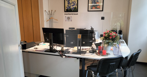 Ein Büro mit einem weißen Tisch, zwei Monitoren und Büromaterial. Im Hintergrund sind Holzschränke und ein Garderobenständer. Auf dem Tisch steht eine Blumenvase mit Blumen und einige Kerzen. An der Wand hängen zwei Bilder mit floralen Motiven.