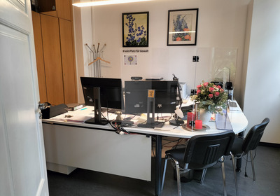 Ein Büro mit einem weißen Tisch, zwei Monitoren und Büromaterial. Im Hintergrund sind Holzschränke und ein Garderobenständer. Auf dem Tisch steht eine Blumenvase mit Blumen und einige Kerzen. An der Wand hängen zwei Bilder mit floralen Motiven.